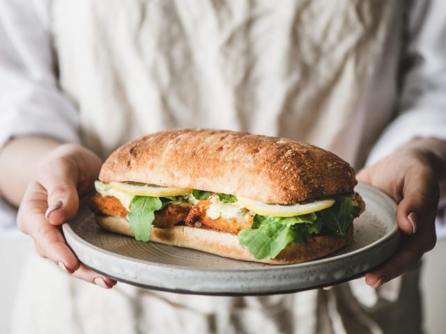Woman in linen apron holding plate with fresh fried fish sandwich with tartare sauce, lemon and arugula, selective focus. Healthy easy breakfast ideas
